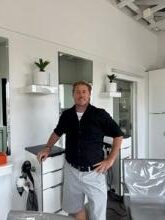 Man in black shirt standing in a modern kitchen with a dog.